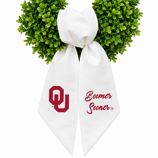 A white bow with long tails, decorated with green boxwood leaves at the top and featuring a red embroidered Oklahoma 'OU' logo on one side and the words 'Boomer Sooner' in red script on the other, shown on a white background.