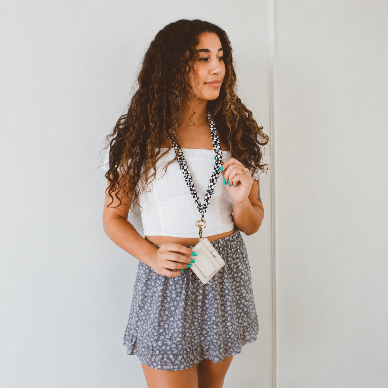 A young woman with long, curly brown hair is wearing a white strapless tube top and a gray floral mini skirt, accessorized with a black and white chaeckered lanyard .