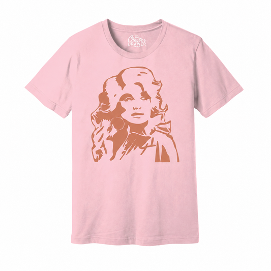 A light pink short-sleeve crewneck t-shirt featuring a stylized graphic print of a woman's face in a warm brownish-orange tone. The brand name 'Cotton's Crafter' is printed inside the collar.