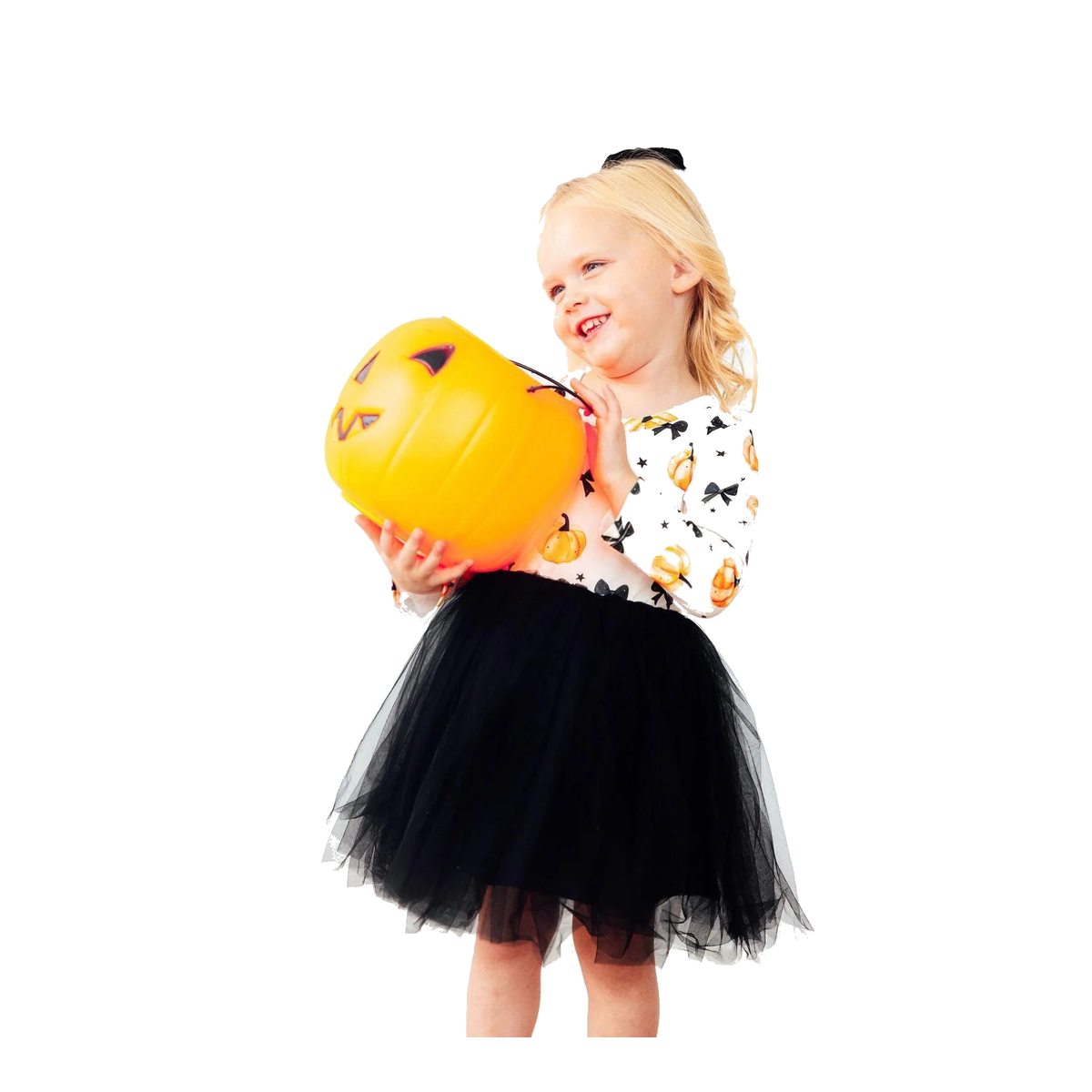 A young girl with blonde hair wearing a black tulle tutu skirt and a white long-sleeve shirt with a black and orange pumpkin print, holding a yellow jack-o'-lantern bucket against a solid white background.