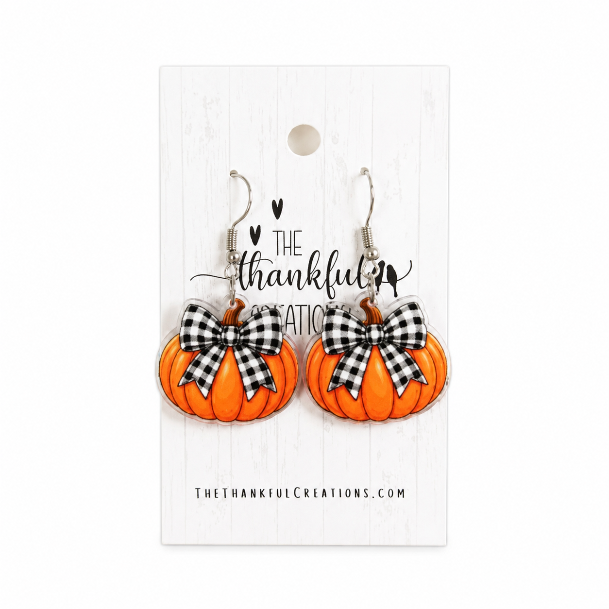 A pair of orange pumpkin-shaped dangle earrings with black-and-white gingham bows, displayed on a white branded card that reads 'The Thankful Creations'.