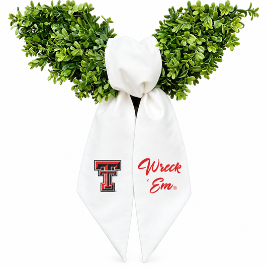 A novelty costume accessory featuring a white satin bow with red and black 'Texas Tech' logo on one side and red cursive 'Wreck 'Em' text on the other, topped with green artificial boxwood branches on a white background.