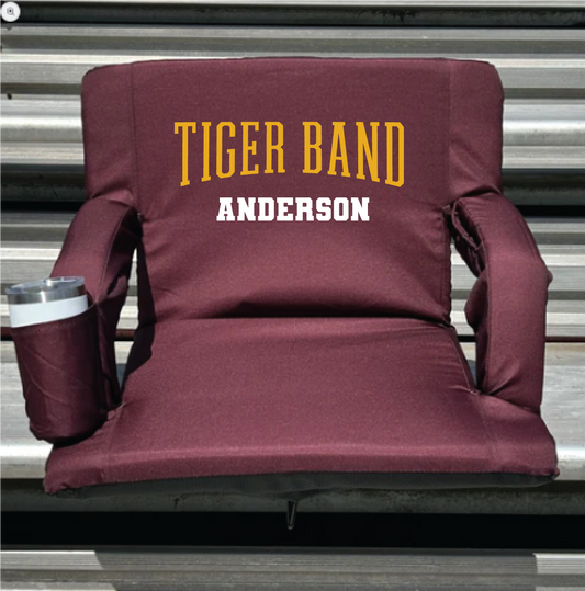 A maroon stadium-style lounge chair with a side cup holder, featuring 'TIGER BAND' in yellow and 'ANDERSON' in white text on the backrest, set against a gray metal bleacher background.